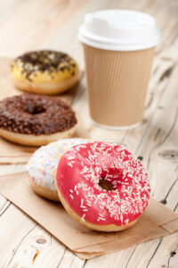 A group of colorful donuts with sprinkles sits on brown parchment paper, with a takeaway coffee cup in the background on a wooden surface. The front donut has pink icing and white sprinkles.