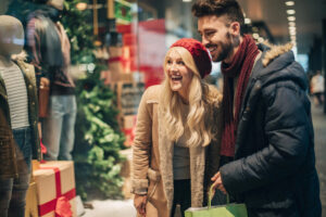 A smiling couple in winter clothing looks at a holiday shop window display decorated with presents and a Christmas tree, appearing happy and excited while shopping.