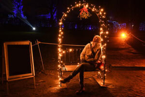 A person in a winter coat and hat sits on a bench decorated with string lights and mistletoe at night, surrounded by festive outdoor lighting and illuminated trees.