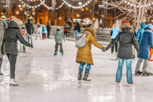 People ice skating outdoors at night, bundled in winter clothing. Many skaters hold hands, and festive string lights hang above, illuminating the scene with a warm, cheerful glow. Trees and buildings are visible in the background.