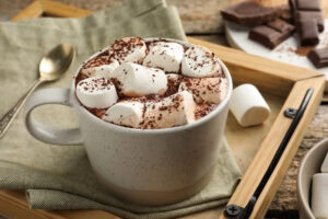 A mug of hot chocolate topped with marshmallows and sprinkled with cocoa powder sits on a green napkin, next to a spoon and pieces of chocolate on a wooden table.