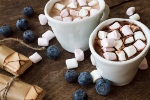 Two white mugs filled with hot chocolate and topped with mini marshmallows sit on a wooden surface, surrounded by fresh blueberries, wrapped wafer cookies, and scattered marshmallows.