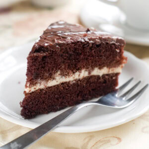 A slice of chocolate layer cake with cream filling and chocolate frosting on a white plate, with a fork beside it and a blurred teacup in the background.