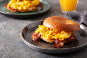 A breakfast sandwich with scrambled eggs and bacon on a toasted bagel sits on a dark plate. Another plate with a similar sandwich and a glass of orange juice are in the background.