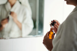 A person in a white bathrobe stands in front of a mirror, holding an amber dropper bottle and drawing liquid from it with a dropper. Their nails are painted black. The background is softly blurred.