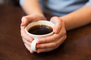 A person holding a white ceramic cup filled with black coffee in both hands, resting on a wooden table. Only the hands and part of the arms are visible.