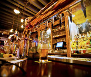 A close-up view of a bar counter with a nearly full pint of beer, shiny brass beer taps, and shelves stocked with bottles of liquor in the background under warm lighting.