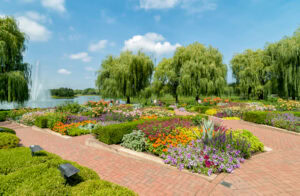 A vibrant botanical garden with colorful flower beds, neatly trimmed bushes, and brick pathways. Willow trees and a large fountain by a pond are visible under a bright blue sky with scattered clouds.