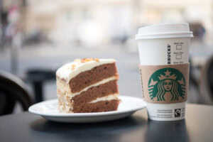 A slice of layered chocolate cake with white frosting sits on a white plate next to a Starbucks takeaway coffee cup on an outdoor café table. The background is blurred.