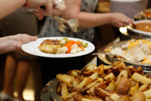 A person holds a plate of food at a buffet, selecting dishes with tongs. The plate contains vegetables, potatoes, and sauce, while a tray of roasted potatoes is visible in the foreground. Other people serve themselves in the background.