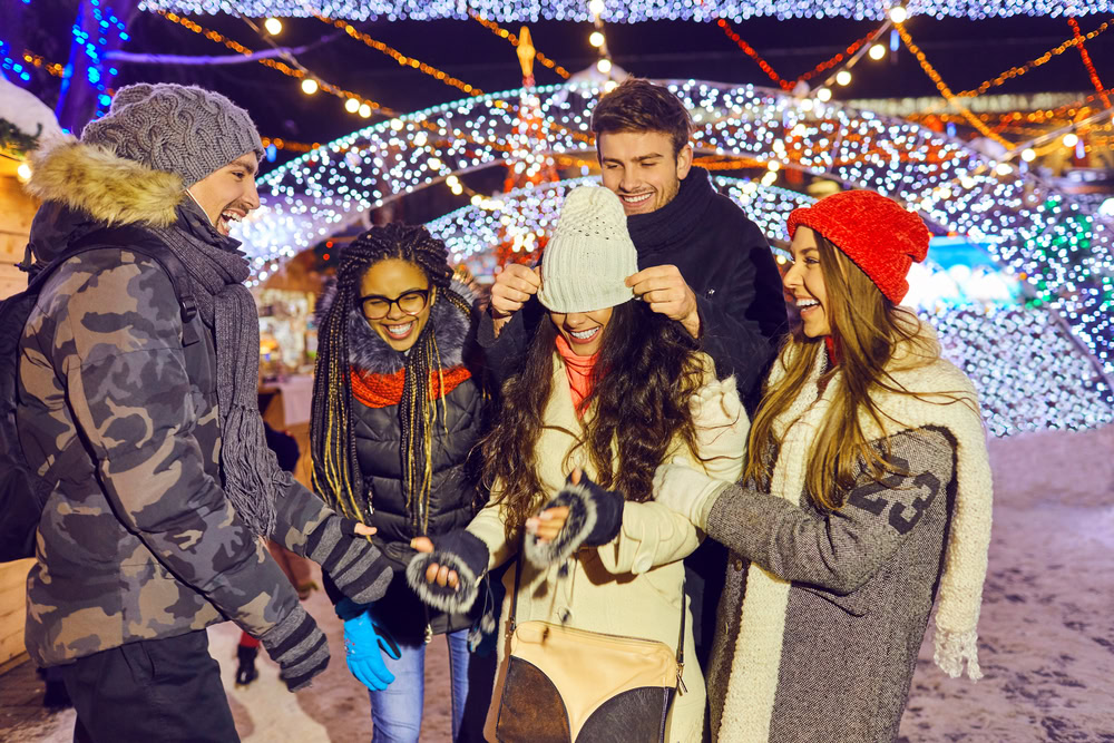 Five friends in winter clothing laugh and smile at a festive outdoor location with holiday lights; one person covers anothers eyes with a hat, creating a playful, joyful atmosphere in the snowy setting.