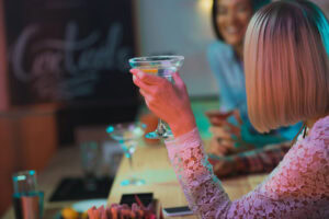 A woman with blonde hair in a white lace top holds a martini glass at a bar, smiling and talking with another woman in the background. The atmosphere is relaxed and colorful, with cocktails on the counter.