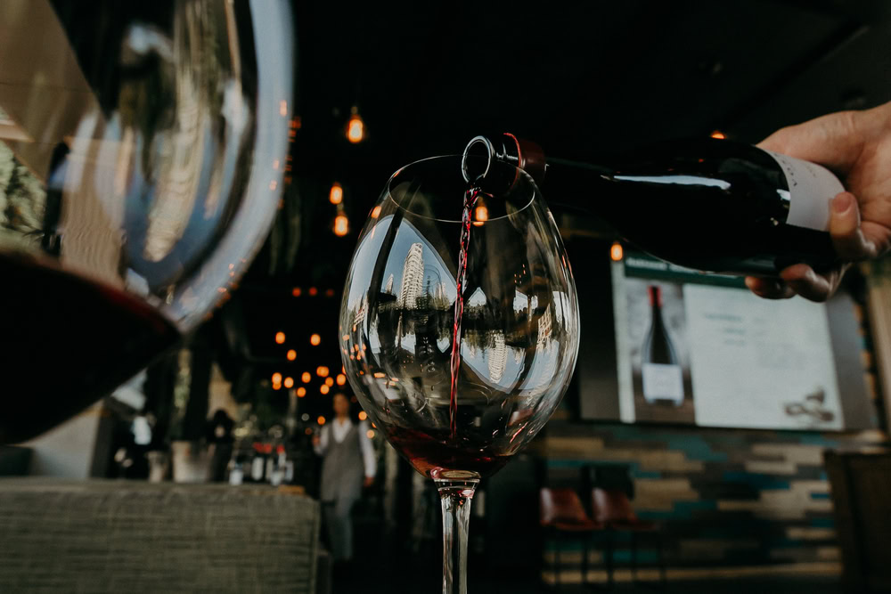 A close-up of red wine being poured into a glass at a stylish restaurant with modern decor and warm ambient lighting in the background.