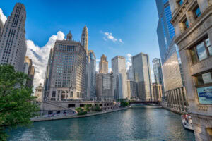 Downtown Chicago skyline with tall buildings lining the Chicago River under a blue sky with clouds. A tour boat is in the river and trees are visible on the riverbank.