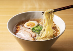 A bowl of ramen with steaming broth, sliced pork, a halved soft-boiled egg, green onions, and noodles being lifted by chopsticks on a wooden table.