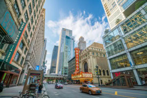 Downtown Chicago street scene with tall buildings, the iconic Chicago Theatre marquee, people walking on the sidewalks, cars on the street, and a bright, partly cloudy sky overhead.