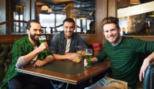 Three smiling men sit together at a pub table, each holding a glass of green beer, with snacks in a bowl in front of them. The setting is cozy and warmly lit, suggesting a festive atmosphere.