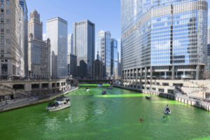 Several boats travel along a bright green river surrounded by tall modern and historic skyscrapers on a sunny day in downtown Chicago.