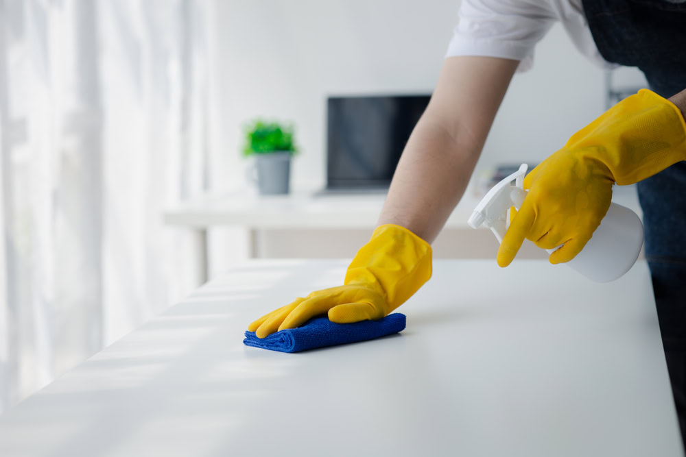 A person wearing yellow rubber gloves cleans a white table with a blue cloth and a spray bottle in a bright room, with a laptop and plant in the background.