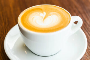 A white cup of cappuccino with heart-shaped latte art on top, placed on a matching saucer on a wooden table.