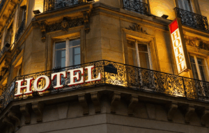 A warmly lit hotel sign on the corner of a stone building with ornate black railings and tall windows, photographed at dusk.