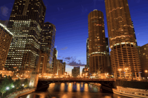 A cityscape of Chicago at dusk shows illuminated skyscrapers and the distinctive Marina City towers along the Chicago River, with lights reflecting on the water and a bridge in the foreground.