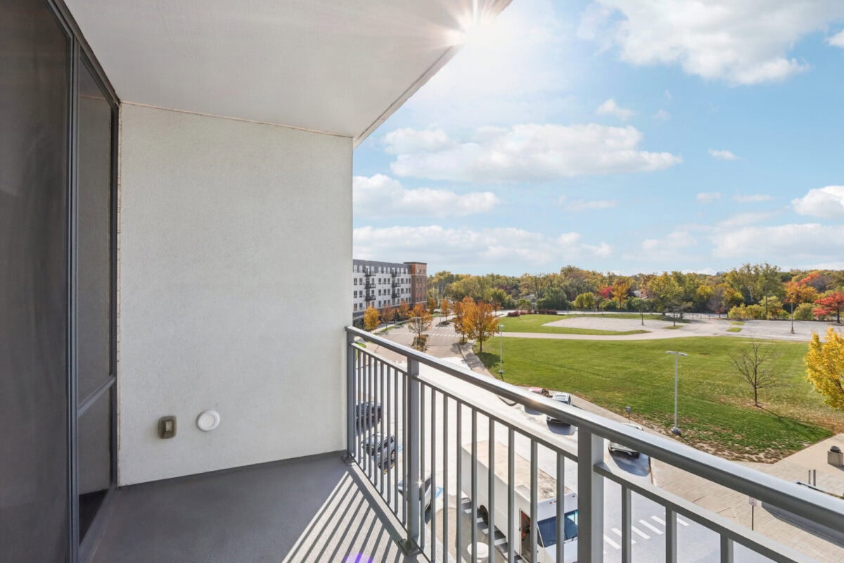 View from a balcony overlooking a green field, parking lot, and a modern apartment building under a bright sky with scattered clouds.