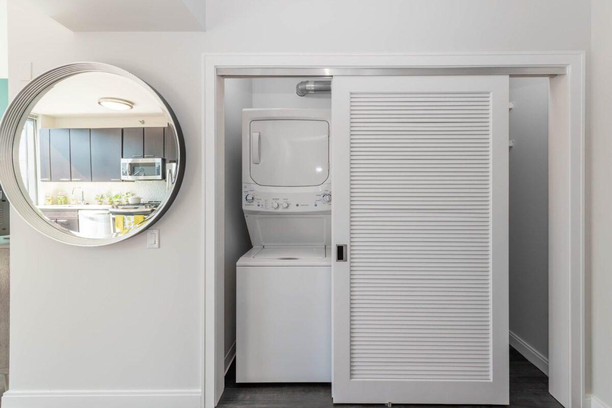 A stacked washer and dryer unit is placed inside a closet with a sliding louvered door, next to a round wall mirror in a modern home interior.