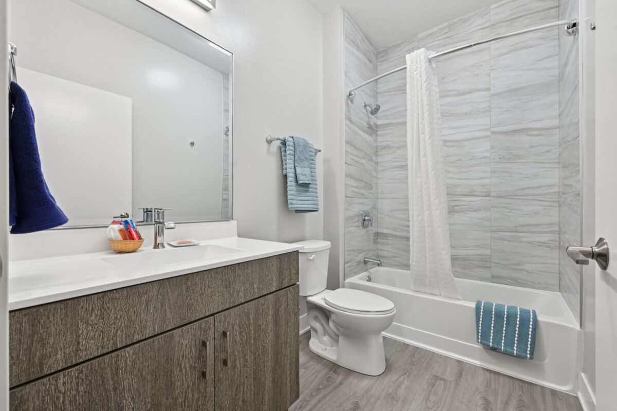Modern bathroom with a wood vanity, white countertop, large mirror, toilet, and a bathtub with a shower curtain and blue towels. Light gray tile and flooring.