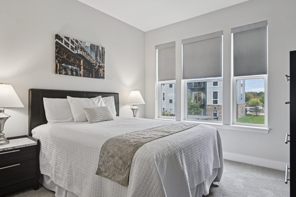 A neatly made bed with white linens is centered between two lamps in a bright bedroom with three large windows and modern decor.