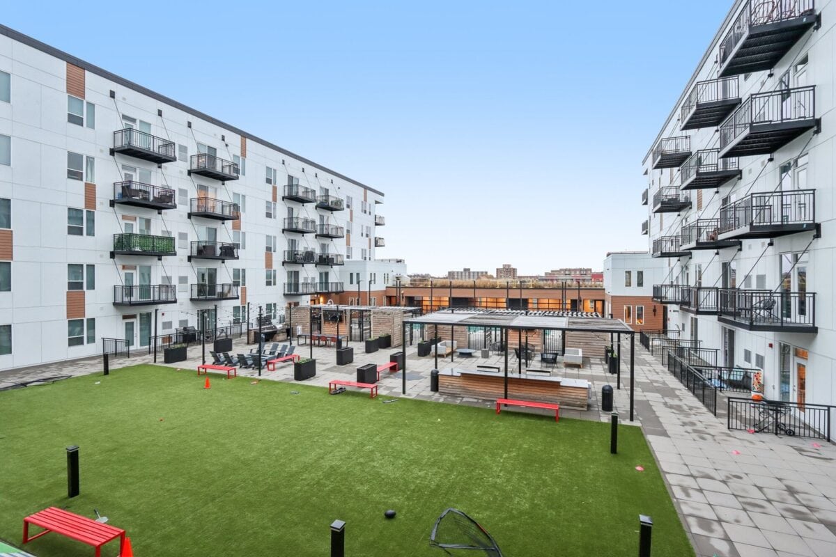 Modern apartment complex courtyard featuring artificial grass, red benches, outdoor seating areas, and balconies on surrounding white buildings under a clear sky.