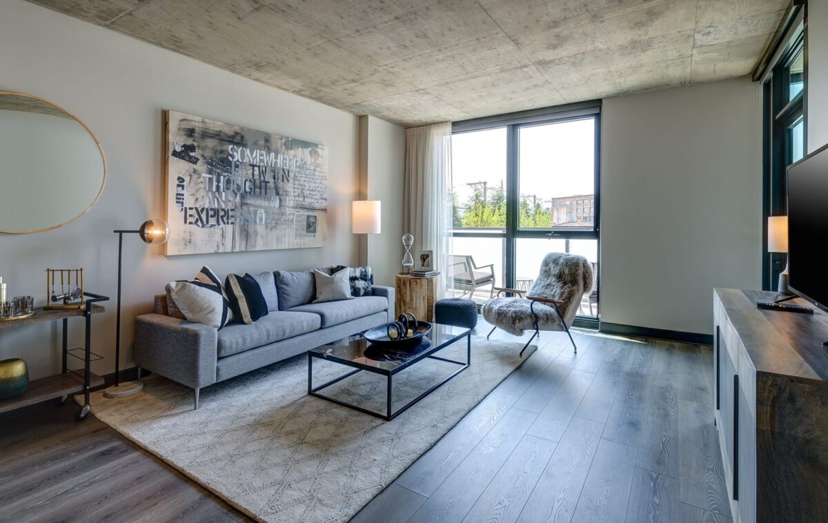 Modern living room with gray sofa, abstract wall art, large window with balcony view, wooden floors, and contemporary furnishings.