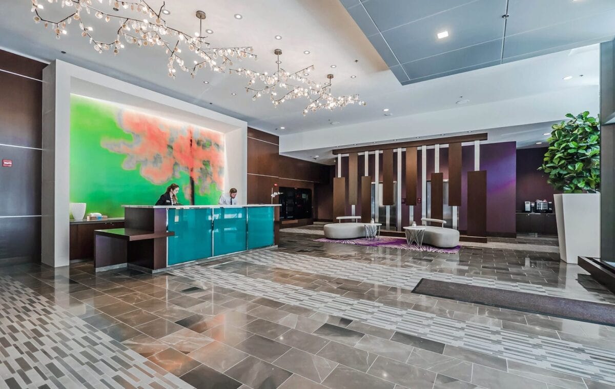 Modern hotel lobby with a tiled floor, green and red abstract wall art, a turquoise reception desk with two staff members, contemporary seating, and decorative lighting.