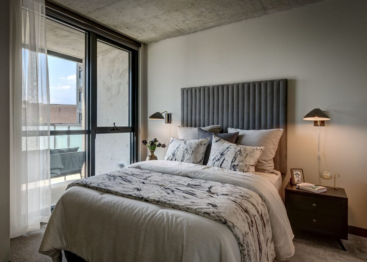 A modern bedroom with a gray upholstered headboard, patterned bedding, two nightstands with lamps, and a glass door leading to a balcony.