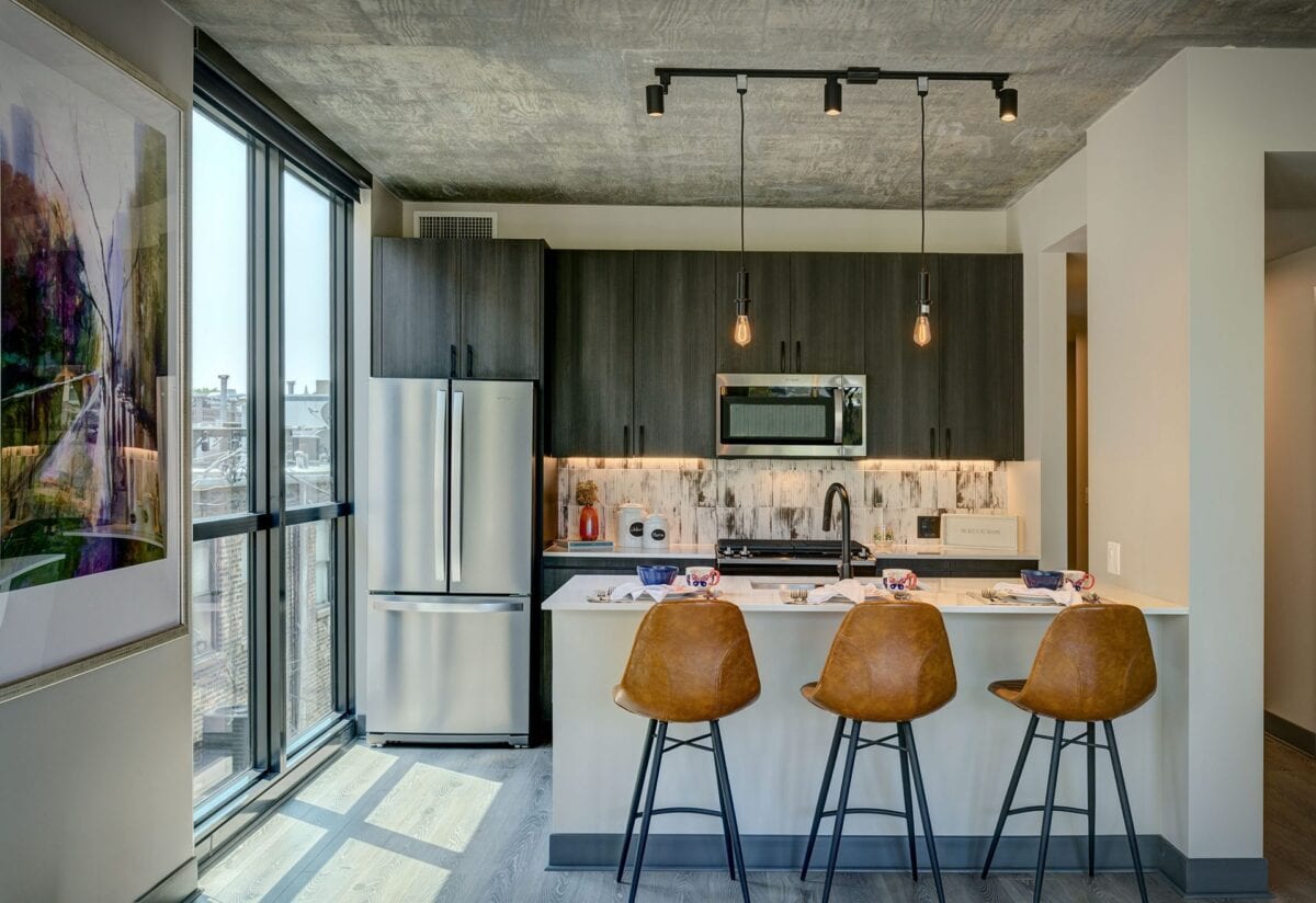 Modern kitchen with dark cabinets, stainless steel appliances, a white island with three brown barstools, pendant lighting, and large window with city view.