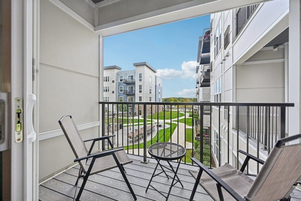 A small balcony with two chairs and a round table overlooks a landscaped courtyard and modern apartment buildings under a clear sky.