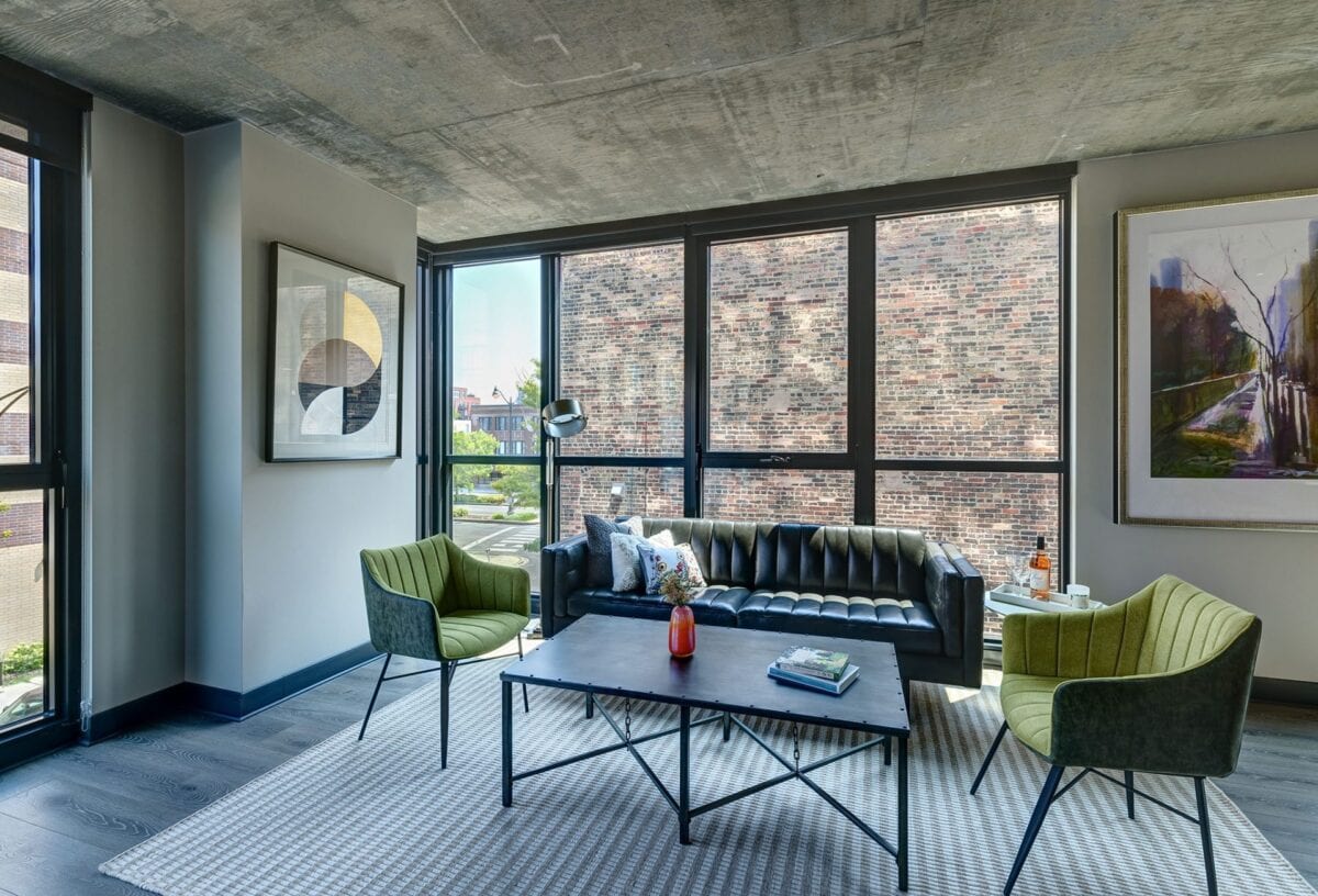 Modern living room with a black leather sofa, two green chairs, a coffee table, large windows, abstract artwork, and neutral decor.