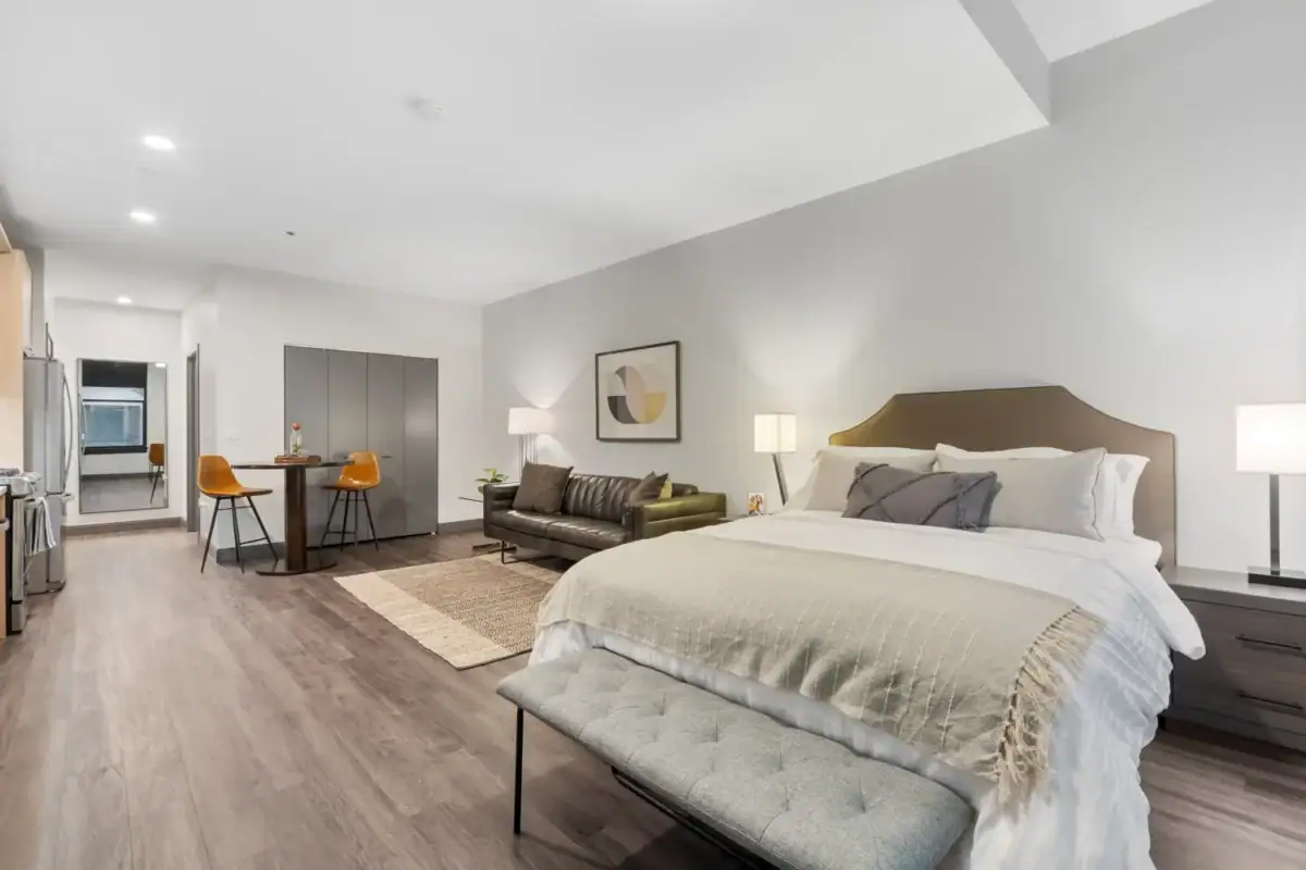 Modern studio apartment featuring a bed, bench, sitting area with a sofa, dining table for two, and kitchen, with neutral decor and wood flooring.