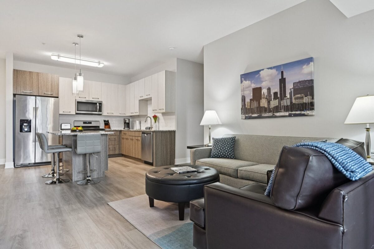 Modern open-concept living room and kitchen with neutral colors, stainless steel appliances, bar stools, and cityscape wall art above the sofa.