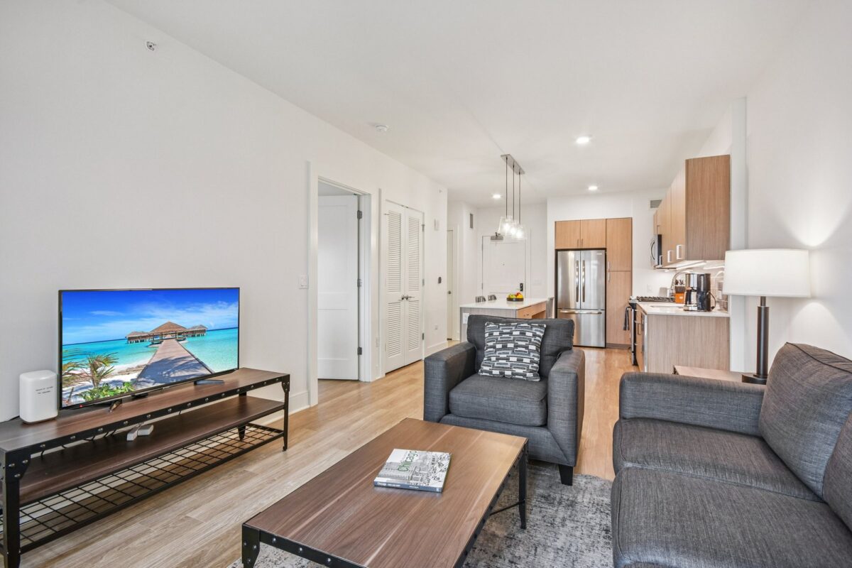 Modern living room and kitchen with neutral decor, gray sofa and chair, flat-screen TV, wood flooring, and stainless steel appliances.
