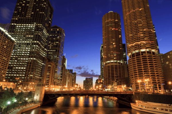 Cityscape at dusk showing high-rise buildings and illuminated lights reflecting on a river, with a bridge spanning across the water.