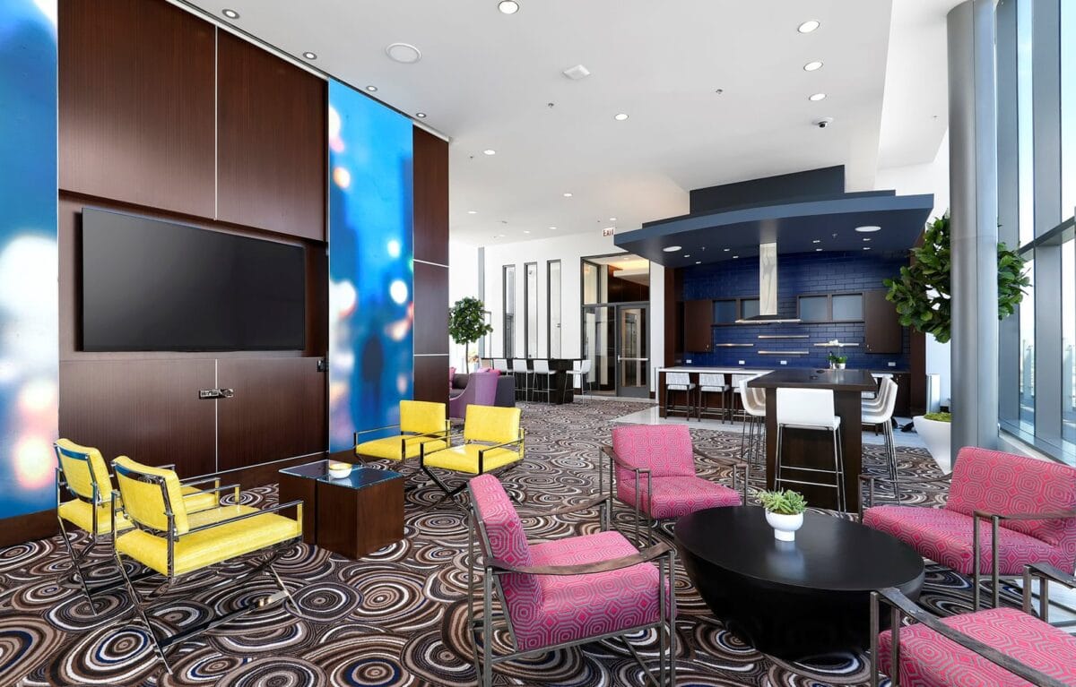 Modern lounge space with patterned carpet, colorful chairs, large TV, small tables with plants, and open kitchen area in the background.