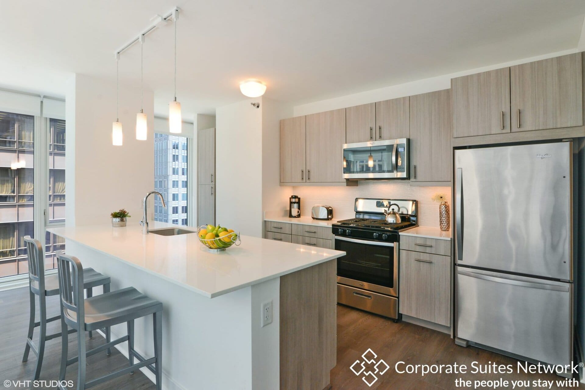 Modern kitchen with light wood cabinets, stainless steel appliances, a center island with barstools, and large windows showing a city view.