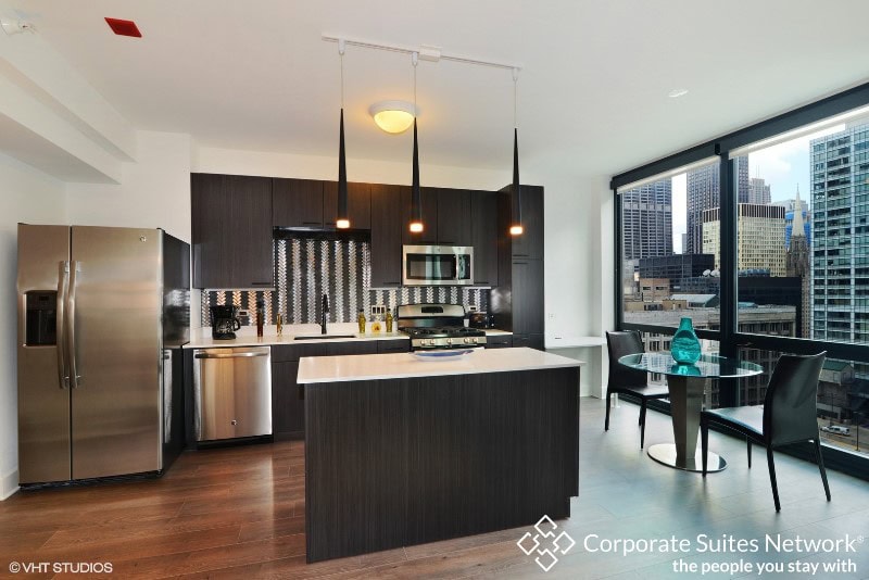 Modern kitchen with dark cabinets, stainless steel appliances, center island, dining table, and large windows showing a city skyline. Corporate Suites Network logo in lower right corner.