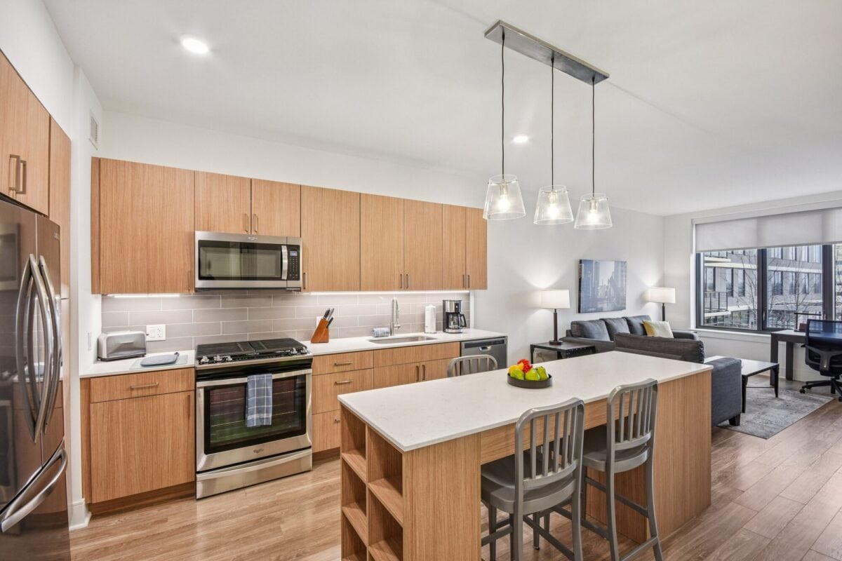 Modern kitchen with wood cabinets, stainless steel appliances, island with three chairs, pendant lights, and an adjacent living area with a sofa, lamps, and large window.