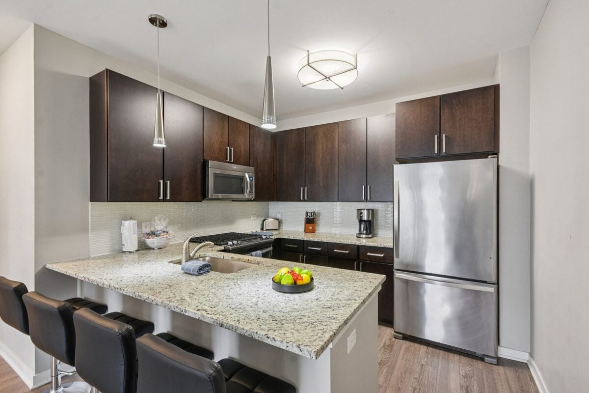 Modern kitchen with dark wood cabinets, stainless steel appliances, granite countertop with a sink, three bar stools, and a bowl of fruit on the counter.