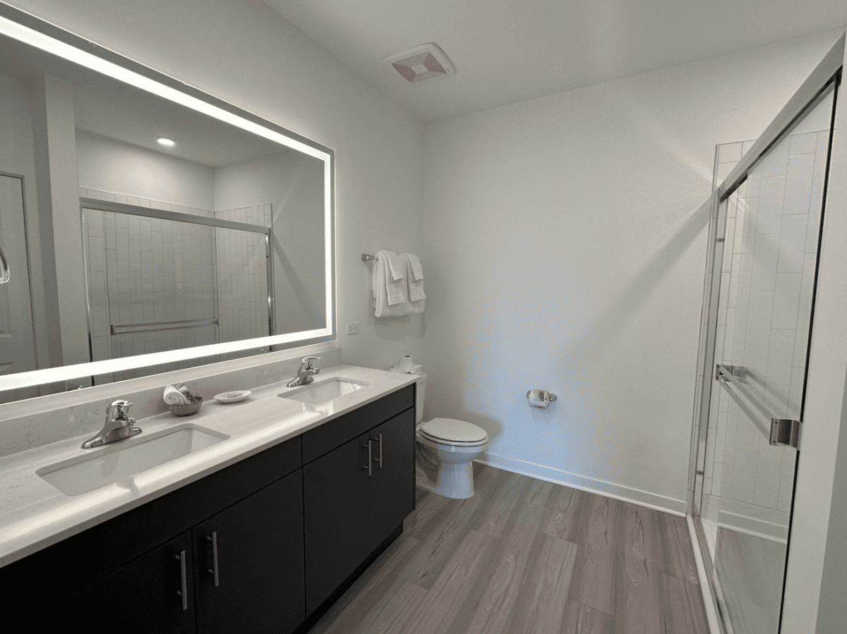 Modern bathroom with double sink vanity, large illuminated mirror, toilet, towel rack, and glass-enclosed shower on wood-look flooring.