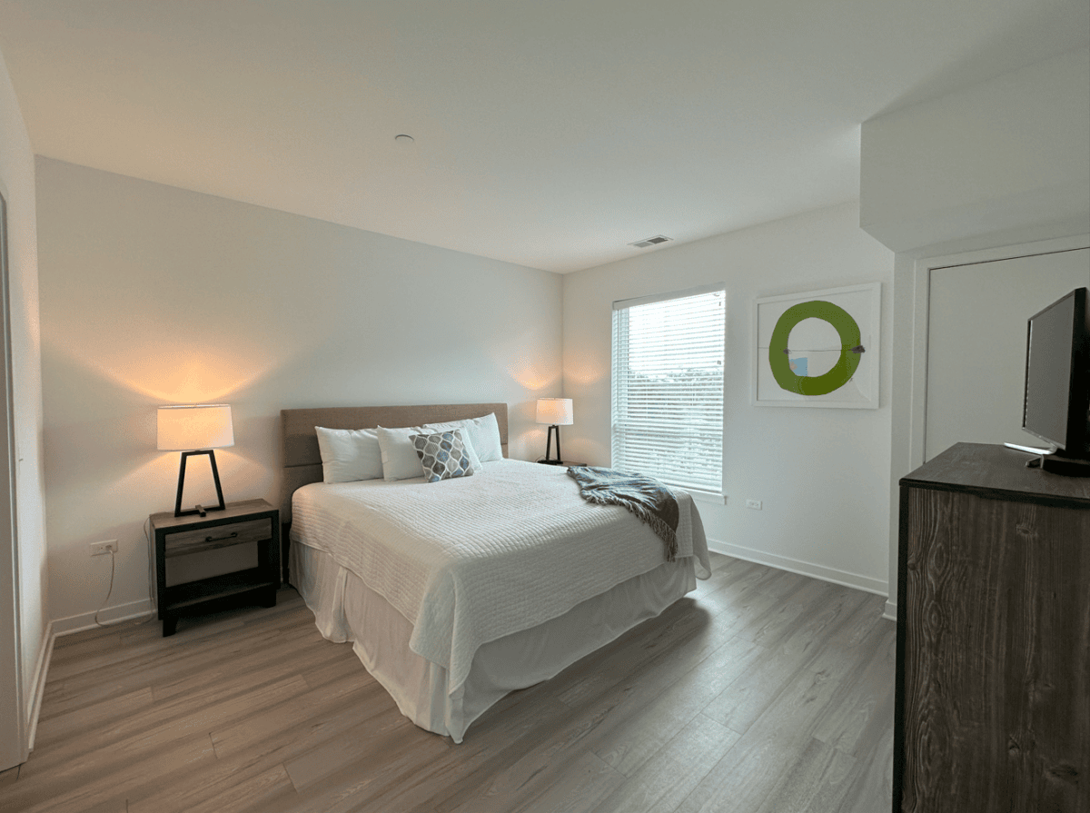 A modern bedroom with a king-sized bed, two bedside tables with lamps, a dresser with a TV, large window, and minimalistic decor.