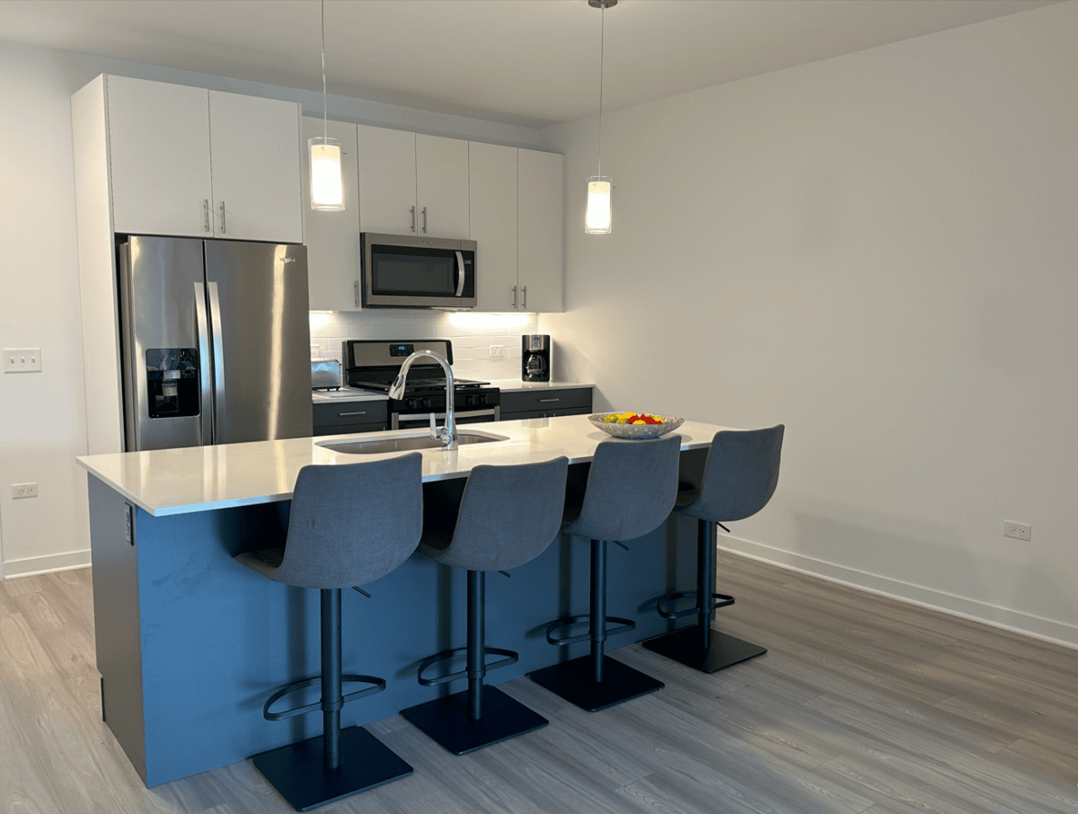Modern kitchen with a large island, four gray barstools, stainless steel appliances, white cabinets, and pendant lights. A bowl of fruit sits on the counter.