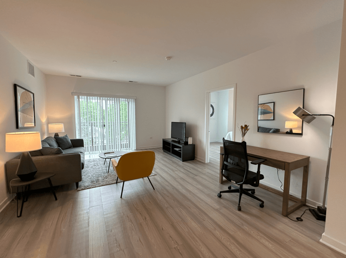 Modern living room with light wood flooring, a sofa, yellow chair, TV, desk with office chair, and sliding glass doors leading to a balcony.